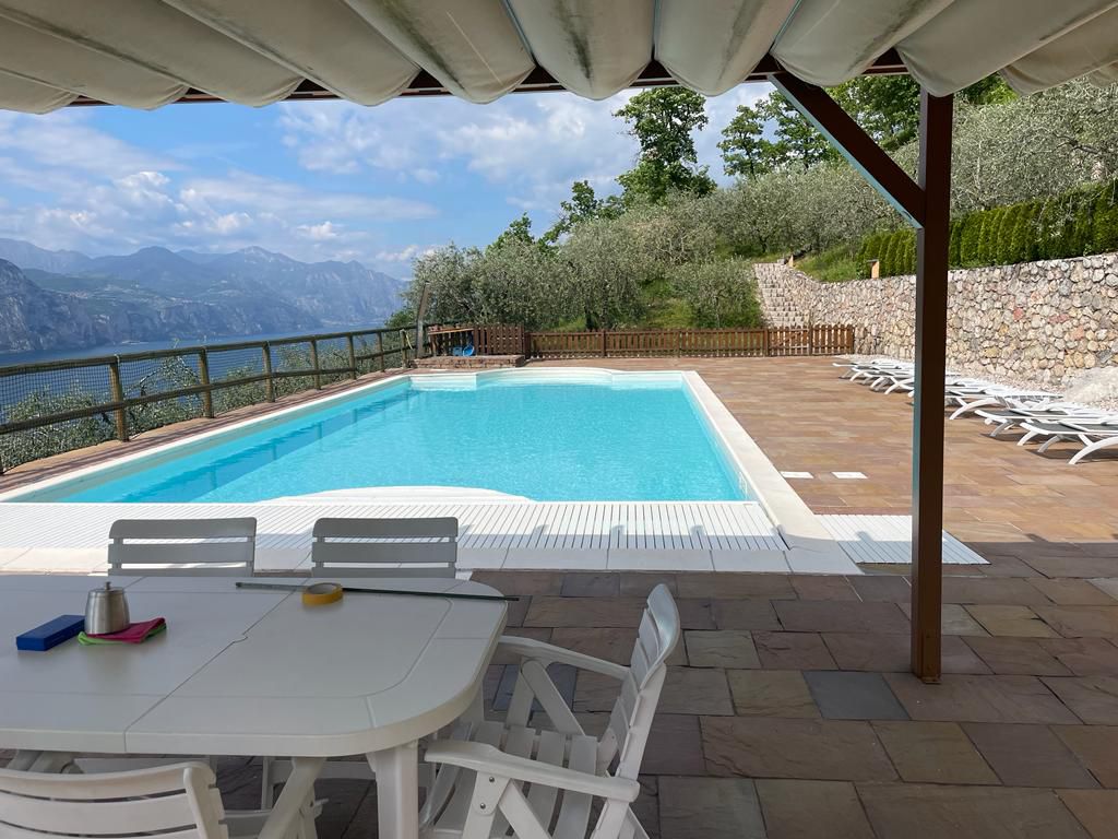 La piscina su un terrazzamento sopra il lago, con pergola, tavolo e lettini, e l’acqua oltre la recinzione.